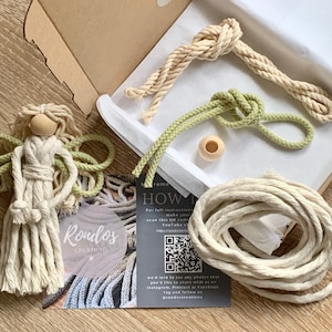 May include: A craft kit for making a macrame angel. The kit includes two spools of rope, one green and one natural, a wooden bead, and a small wooden doll with a natural coloured head and green hair. The kit also includes a card with the company logo, "Rondos Creations", and a QR code for instructions.