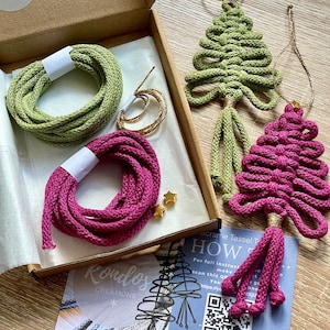 May include: A macrame craft kit featuring green and magenta cords, gold star beads, and two finished Christmas tree ornaments. The kit includes instructions and is packaged in a cardboard box. The cords are for making macrame tassels.
