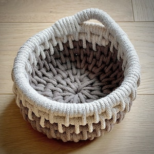 May include: A brown and white crocheted basket with a handle. The basket is made of thick rope and has a circular shape.