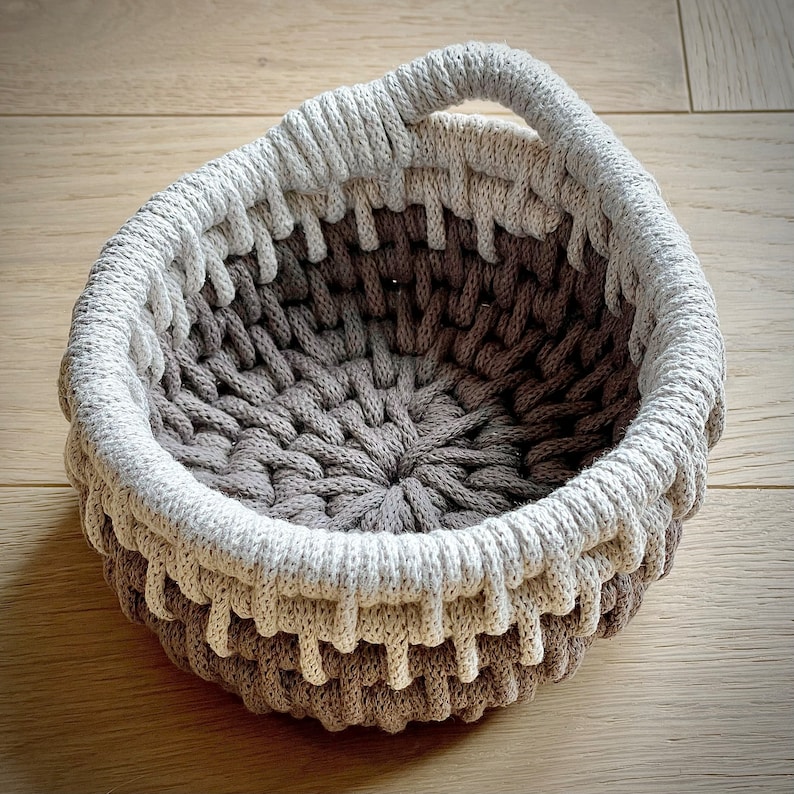 May include: A brown and white crocheted basket with a handle. The basket is made of thick rope and has a circular shape.