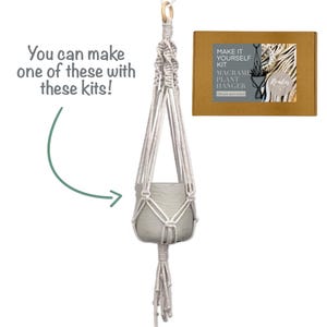 May include: A macrame plant hanger kit featuring a beige cord hanger with a wooden ring at the top and a grey pot inside. The kit includes a brown cardboard box labelled 'MAKE IT YOURSELF KIT MACRAME PLANT HANGER' and text stating 'You can make one of these with these kits!'