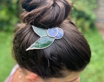 May include: A stained glass hair clip with green leaves and blue berries. The clip is worn in a woman's hair.