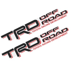 Op de afbeelding: Twee identieke stickers met de tekst TRD OFF ROAD in dikke zwarte letters met rode omlijningen. Onder de hoofdtekst staan de woorden TOYOTA RACING DEVELOPMENT in kleinere rode hoofdletters op een witte achtergrond.