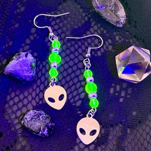 May include: A pair of silver alien earrings with green beads and a silver alien head charm. The earrings are displayed on a black lace surface with iridescent green sequins and crystals.