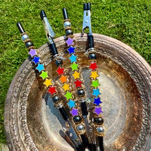 May include: Four black alligator clips with rainbow star charms and decorative beads. The clips are arranged on a silver platter, with a green grassy background. The star charms are in a rainbow of colors.