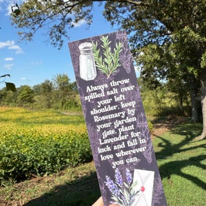 May include: A bookmark with a dark purple background featuring illustrations of a salt shaker, rosemary, lavender, and an envelope. The text reads: "Always throw spilled salt over your left shoulder..." The bookmark is held in front of a field and trees.
