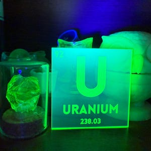 May include: A glowing green Uranium sign with the element symbol 'U' and atomic weight 238.03. A glowing green rock is in a glass jar. An owl-shaped object is in the background.
