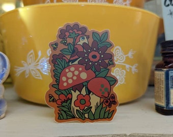 May include: A colorful sticker featuring a whimsical design of mushrooms, flowers, a bird, bees, and a butterfly. The sticker has a brown background and is surrounded by a yellow bowl and other vintage items.