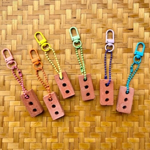 May include: Six miniature brick keychains in various colors, including pink, orange, yellow, green, purple, and blue. Each brick has three holes and is attached to a chain and a clasp. The keychains are arranged on a woven, light brown surface.