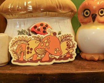 May include: A vintage-style sticker featuring a group of mushrooms with brown stems and orange caps. The mushrooms are surrounded by green leaves and a snail is perched on top of one of the mushrooms.