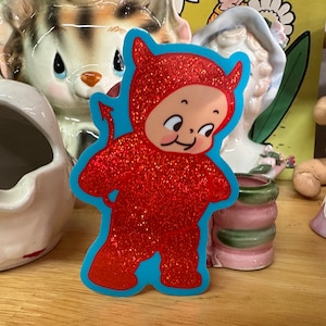 May include: A red glitter devil sticker with a blue background. The devil is wearing a red suit with a tail and horns. The devil has a mischievous smile on its face.