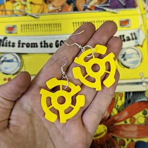 May include: A pair of bright yellow earrings shaped like 45 RPM record adapters. The earrings are held in a hand against a colorful background with a yellow van and the text "Hits from the 60s Hall".