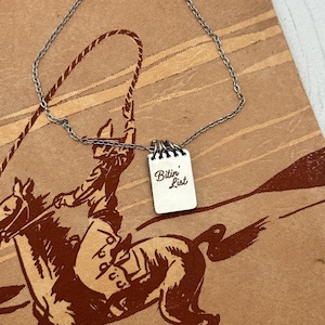 May include: A silver-colored necklace with a small, rectangular pendant. The pendant has the words "Bitin' List" in a handwritten style. The necklace is displayed on a vintage-style brown background with a cowboy illustration.