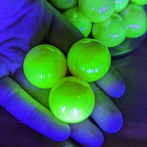 May include: Bright green, glowing spheres are held in a hand, with more spheres visible in a glass container. The spheres have a smooth, reflective surface. The scene is illuminated with a blue light, creating a striking contrast.