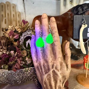 May include: A pair of neon green teardrop earrings with silver hooks. The earrings are displayed on a sculpted hand statue. The background includes a decorative floral arrangement and a horse statue.