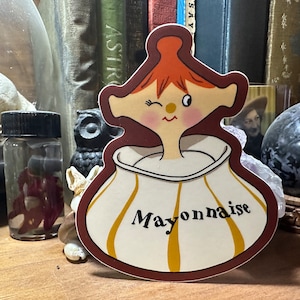 May include: A cartoon sticker of a winking girl with red hair wearing a white and yellow striped dress with the word "Mayonnaise" printed on it.