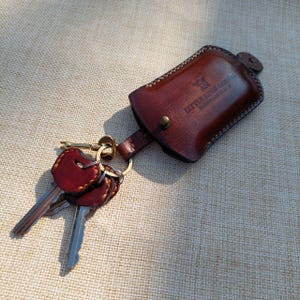 Puede incluir: Un llavero de cuero marrón con el texto "LITTLE KING GOODS" en relieve. El llavero está unido a un llavero con varias llaves y un llavero decorativo de cuero rojo.