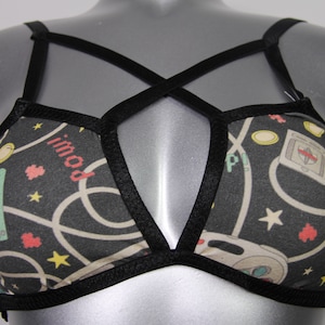 Könnte beinhalten: Schwarzer BH mit einem Gaming-Muster mit Spielkonsolen, Sternen, Herzen und dem Wort "imod". Der BH hat schwarze Träger und Besätze und wird auf einer Schaufensterpuppe präsentiert.