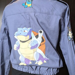 May include: A blue denim jacket with a large cartoon image of a blue and purple turtle Pokemon on the back. There are two smaller images of the same Pokemon on the shoulders.