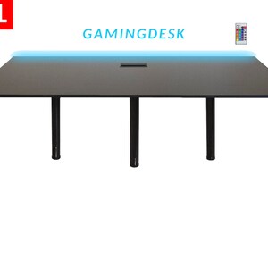 Peut inclure: Un bureau de jeu noir avec une bande lumineuse LED bleue le long du bord avant. Le bureau a quatre pieds noirs et une découpe pour la gestion des câbles. Le texte "GAMINGDESK" est imprimé sur le bureau.