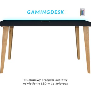Puede incluir: Escritorio de juego negro con patas de madera y un sistema de gestión de cables integrado. El escritorio tiene iluminación LED y el texto "GAMINGDESK" se muestra encima del escritorio. También se ve un mando a distancia.