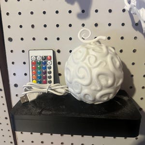 May include: A white, spherical decorative light with a swirled pattern, resembling a fruit. It sits on a black shelf with a remote control and USB cable. The remote has color options. The light is a unique home decor item.