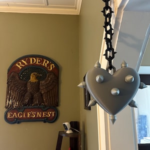 Può includere: Un oggetto grigio a forma di cuore con punte color argento che sporgono dalla superficie, sospeso a una catena di plastica nera. Il cuore presenta una finitura liscia e opaca ed è fissato alla catena da un piccolo anello grigio in alto.