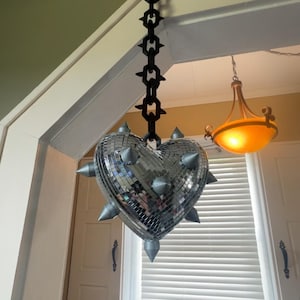 May include: A heart-shaped disco ball covered in small silver mirrored tiles, featuring several protruding silver spikes. The heart is suspended from a black plastic chain with a spiked design, hanging vertically from a door frame.