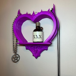May include: A purple heart-shaped shelf with a small brown bottle labeled "XXX" on the shelf. A silver chain with a pentagram charm hangs on the left, and a black cord with a decorative bead hangs on the right.