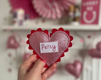 Könnte beinhalten: Ein pinkfarbener, glitzernder herzförmiger Anhänger mit rotem, gezacktem Rand und dem Namen "Percy" auf einem weißen Rechteck gestickt. Im Hintergrund befinden sich Valentinstagsdekorationen.