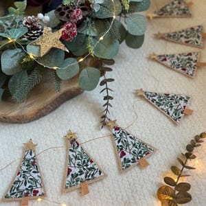 Puede incluir: Una guirnalda festiva de árboles de Navidad con un estampado floral de hojas verdes y bayas rojas. Cada árbol está coronado con una estrella dorada con purpurina y tiene una base de madera. La guirnalda está ensartada en un hilo dorado fino.