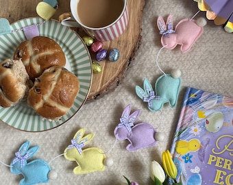 May include: A charming Easter still life with a cup of tea, hot cross buns, and a garland of felt bunnies. A book with a floral cover and a bouquet of tulips add to the festive scene. Chocolate eggs are scattered around the plate and garland.