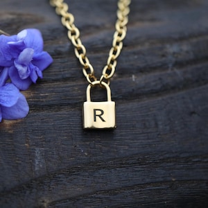Pode incluir: Um colar de corrente de ouro com um pingente de cadeado. O cadeado tem a letra "R" gravada.