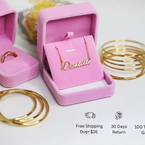 May include: Two pink velvet jewelry boxes, one with a gold ring and the other with a gold necklace that says "Danielle". There are also three gold bangles stacked on top of each other.