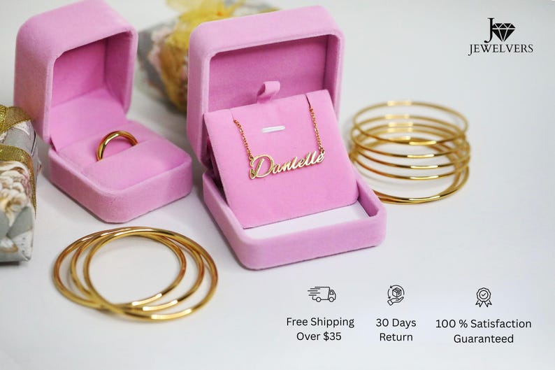 May include: Two pink velvet jewelry boxes, one with a gold ring and the other with a gold necklace that says "Danielle". There are also three gold bangles stacked on top of each other.