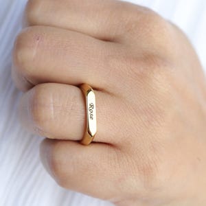 May include: A gold-colored ring with a rectangular band engraved with the name "Rose".