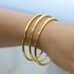 May include: Three gold bangle bracelets stacked on a wrist. The bracelets are engraved with the words "And Your Heart", "Charlotte", and "Always".