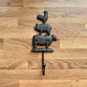 May include: A cast iron hook featuring a rooster, pig, and cow design. The hook is black and has a rustic appearance.