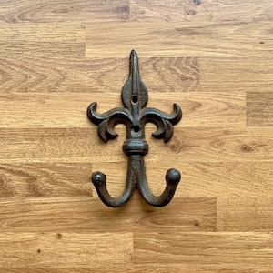 May include: A black cast iron double hook with a decorative fleur-de-lis design. The hook is mounted on a wooden surface.