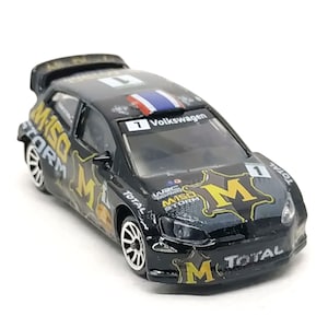 May include: Black Volkswagen Polo WRC race car with a yellow and black MISO STORM logo and TOTAL sponsor decals. The car has a number 1 on the side and the windshield.
