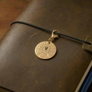 Puede incluir: Un charm redondo dorado con un diseño de mapa y un alfiler de ubicación, sujeto a un cordón negro. El charm está sobre un diario de cuero marrón, lo que sugiere un tema de viaje o de recuerdos.