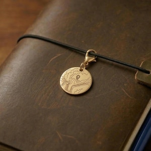 May include: A gold-colored charm with a detailed map design hangs from a black cord, attached to a brown leather journal. The charm is circular and features etched lines. The journal is closed, with a pen visible.