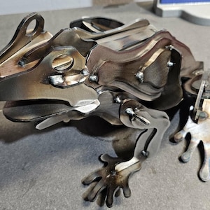 May include: A metal frog sculpture with a layered design. The sculpture is made of welded metal pieces, showcasing a detailed frog form with visible welds and a natural, metallic finish. The frog has outstretched legs and a detailed head.