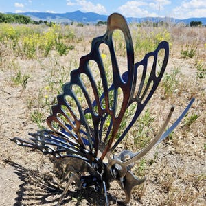 Puede incluir: Escultura de mariposa metálica con intrincados cortes en las alas, mostrando un degradado de tonos bronce, azul y plata. La obra de arte está situada al aire libre, en un campo, con un cielo azul y nubes en el fondo.