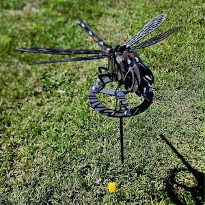 Pode incluir: Uma escultura de lib&eacute;lula em metal com partes do corpo escuras e segmentadas e asas intrincadas e vazadas. O inseto &eacute; montado sobre uma base circular de metal sustentada por uma estaca vertical fina, criando um ornamento de jardim tridimensional.