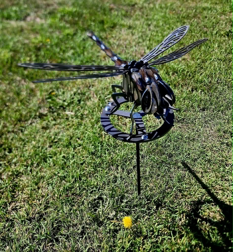 Pode incluir: Uma escultura de lib&eacute;lula em metal com partes do corpo escuras e segmentadas e asas intrincadas e vazadas. O inseto &eacute; montado sobre uma base circular de metal sustentada por uma estaca vertical fina, criando um ornamento de jardim tridimensional.