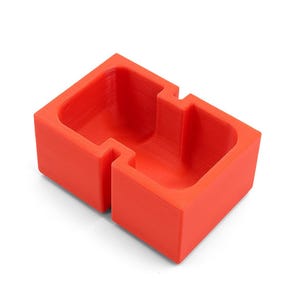 Könnte beinhalten: Ein leuchtend roter, 3D-gedruckter Behälter mit zwei Fächern. Die rechteckige Form hat ein abgestuftes Design, wobei jedes Fach einen vertieften Innenraum aufweist. Das Objekt besteht aus massivem Material und steht auf einer weißen Oberfläche.