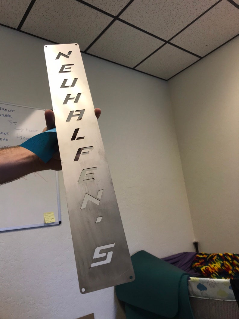 May include: A silver metal sign with the word "NEHI" cut out in a stylized font.