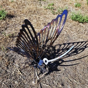 Könnte beinhalten: Eine Metall-Schmetterlingsskulptur mit filigranen Flügelausschnitten. Die Flügel zeigen einen Farbverlauf von Bronze bis Blau. Die Skulptur steht auf einem Bett aus trockenem Gras und Erde und wirft einen Schatten.