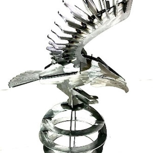 Puede incluir: Una escultura de metal plateado de un águila con las alas extendidas, posada sobre una esfera. El águila está detallada con intrincados recortes y la esfera está hecha de múltiples anillos.